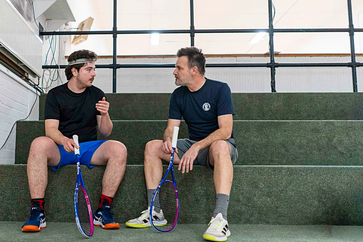 Two men sitting courtside talking with Vadetep squash rackets